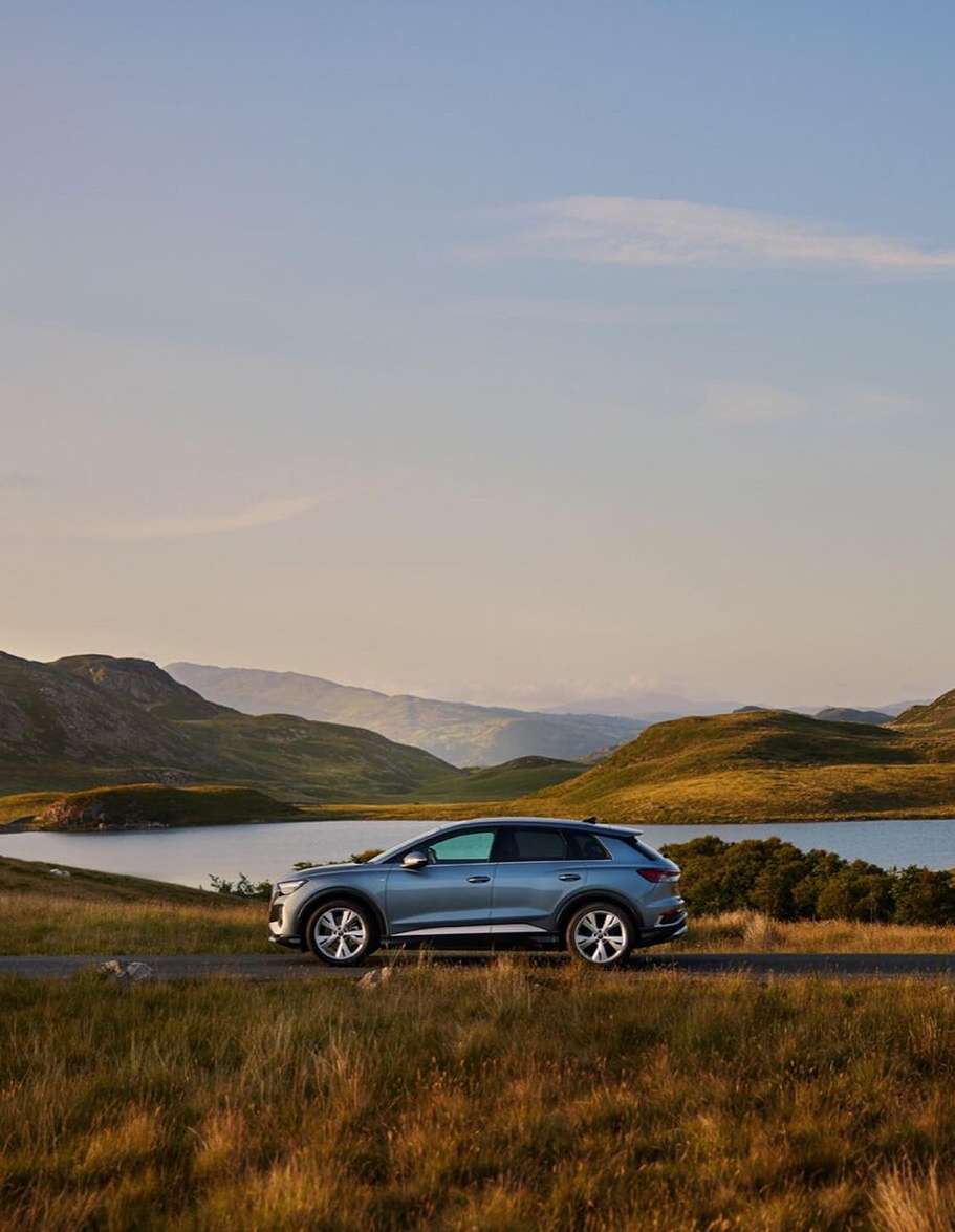 Silver car drives on a rural road by a lake with rolling hills under a blue sky during daytime, enhancing its scenic beauty.