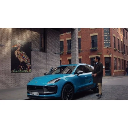 Turquoise Porsche Macan parked on a brick street with a stylish man standing nearby, set against a backdrop of brick buildings and a mural.