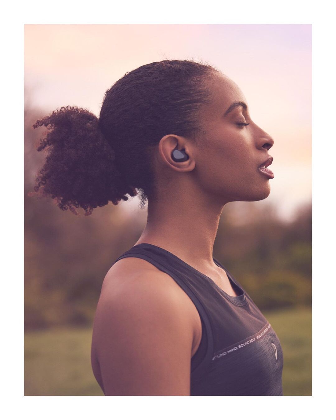 Profile view of an African-American woman with an earbud in her ear, eyes closed, and athletic wear, in a grassy outdoor setting.