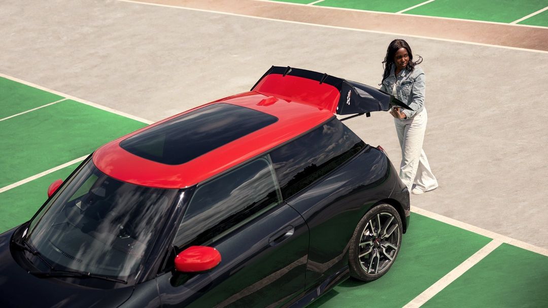 Black and red convertible car is parked on a concrete surface with green lines, with a smiling woman standing near the back of it.