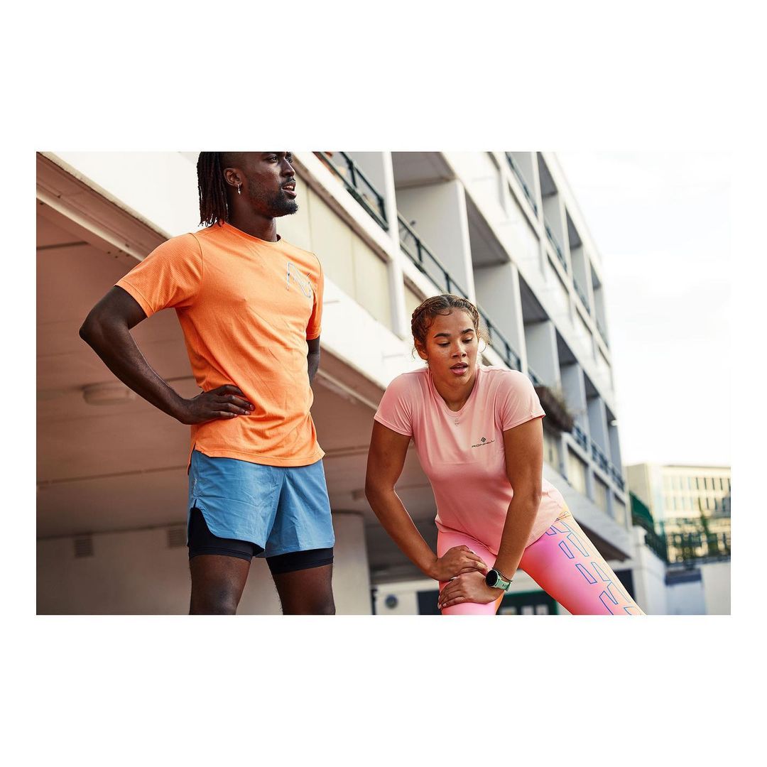 A man and woman in athletic wear are outside on a track near a building. The woman is stretching her leg, while the man stands nearby.