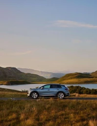 Silver SUV parked on a paved road beside a tranquil lake with rolling green hills and mountains under a clear sky
