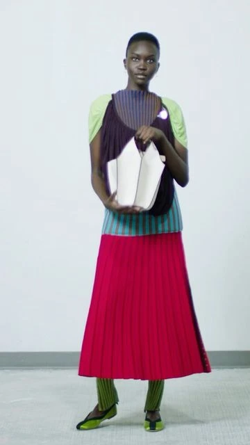 A Black female model poses in a photo studio wearing a colorful, layered outfit consisting of skirt, top, leggings, and flats, holding a handbag.