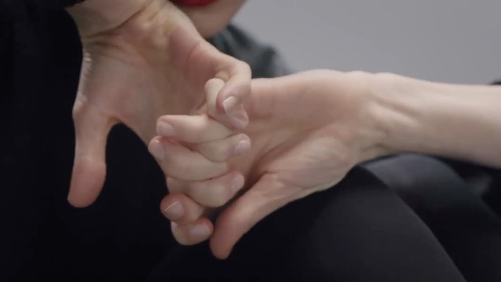 Close-up of hands gesturing, forming a shape; fair skin, red lipstick, grey top and black clothing visible against a neutral background.