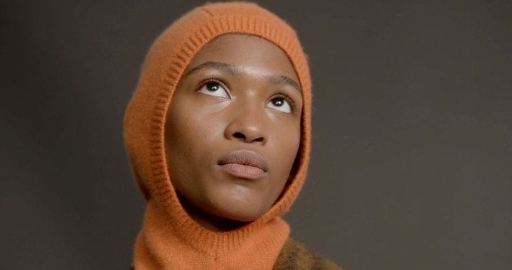 Close-up of Black female looking up in orange balaclava against grey background. Beauty shot, fashion, skincare, studio lighting.