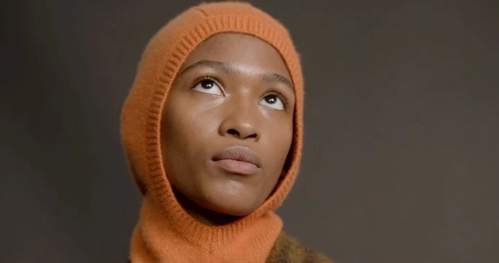 Close-up of Black female looking up in orange balaclava against grey background. Beauty shot, fashion, skincare, studio lighting.