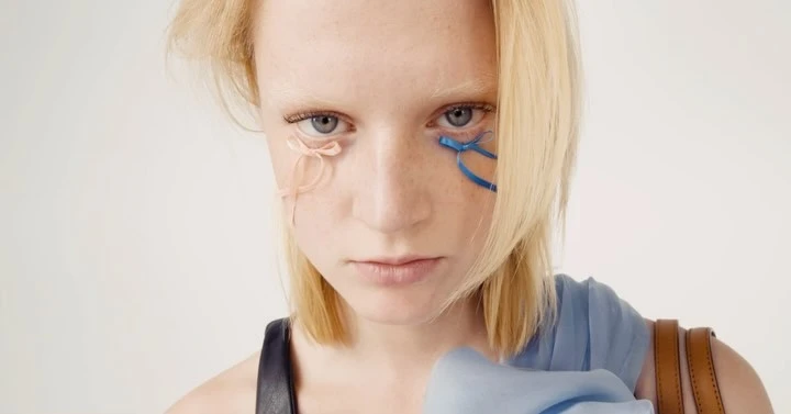 Close-up of a model with blonde hair and freckles, adorned with pink and blue ribbons near the eyes, against a plain background.