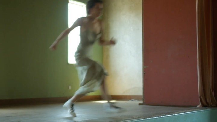 A woman with dark hair sprints across a stage in a long dress, captured indoors with natural lighting and blurred motion in the scene.