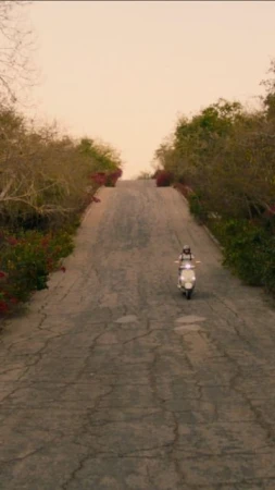 A woman rides a white scooter up a cracked asphalt road lined with green trees and red flowers on a sunny day with a hazy sky.