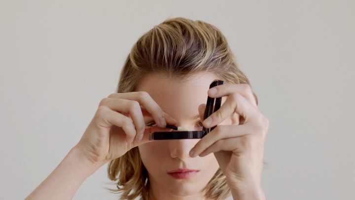 Adult woman holds an open makeup compact with tweezers in an indoor studio setting. The woman is fair skinned with blonde hair.