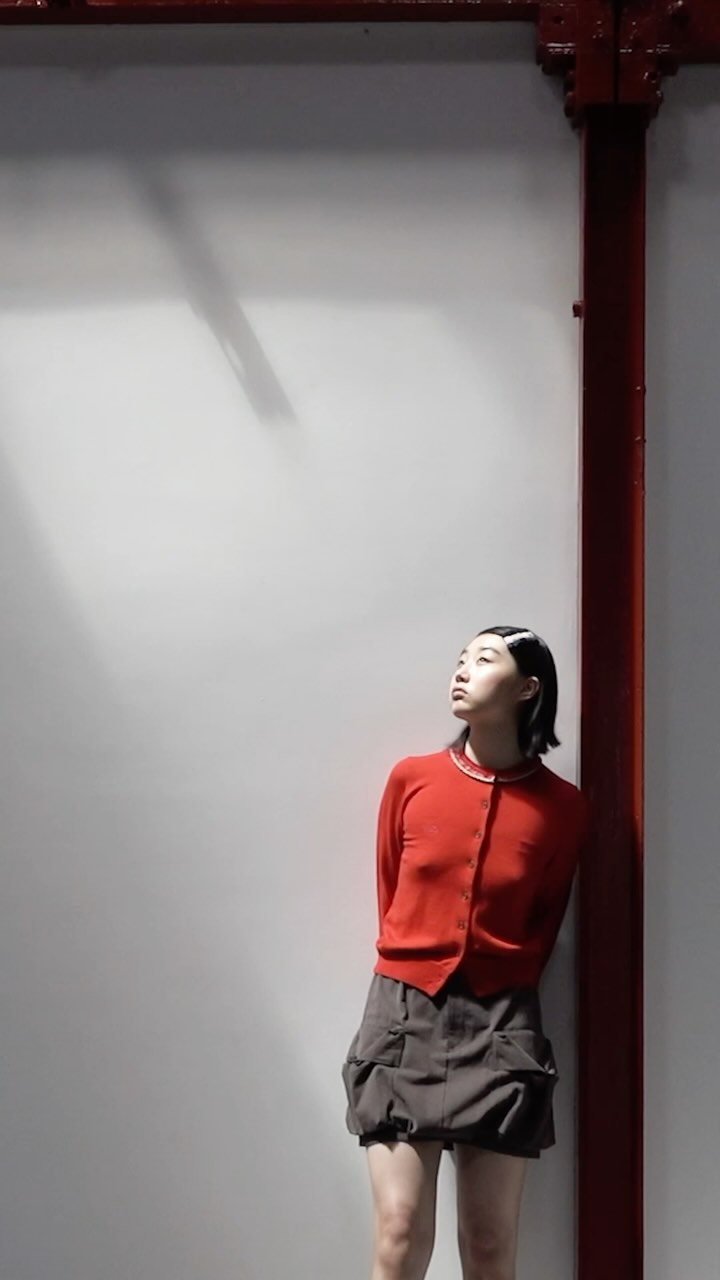 Young woman in red cardigan and grey skirt stands by a white wall with a red support beam looking up in an indoor environment.