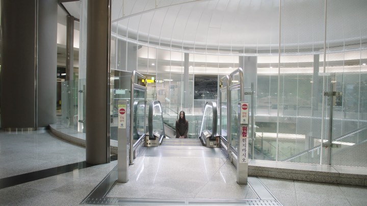 A young woman stands in front of a pair of escalators in a modern, brightly lit, spacious building with metallic and glass design elements.