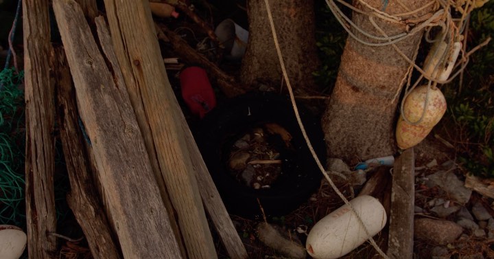 A collection of weathered wooden planks and discarded items such as rope, plastic bottles, and buoys, set against the backdrop of tree trunks.