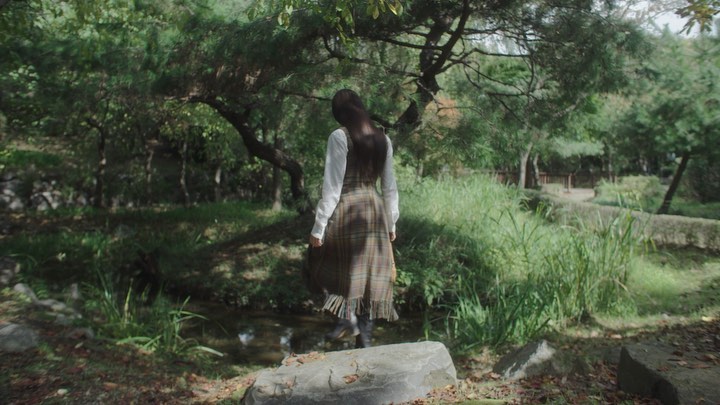 A woman with long hair walks across a small stream in a lush green park wearing a plaid dress and boots in a scenic outdoor setting.