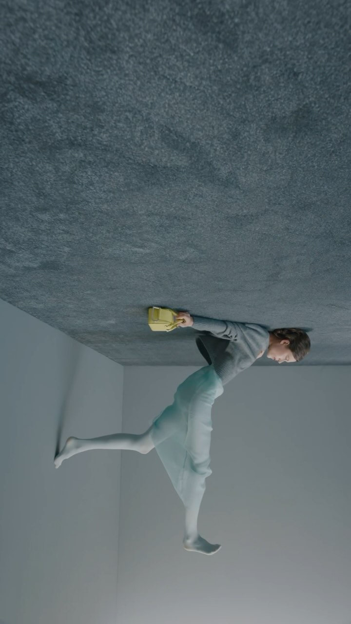 Conceptual fashion shot of a young adult female in a grey top and sheer skirt, holding a yellow purse in a grey indoor setting.