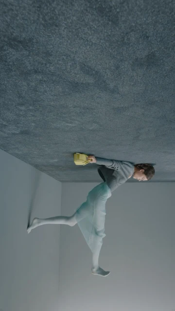 Conceptual fashion shot of a young adult female in a grey top and sheer skirt, holding a yellow purse in a grey indoor setting.
