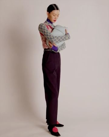 A stylish woman in a patterned sweater and dark pants holds a blue purse while standing in front of a plain white background.