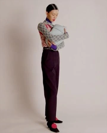 A stylish woman in a patterned sweater and dark pants holds a blue purse while standing in front of a plain white background.