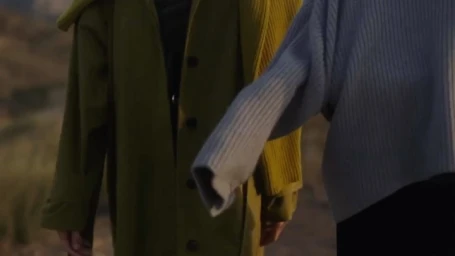 Two people outdoors, one wearing an olive green coat and the other in a light blue ribbed sweater.