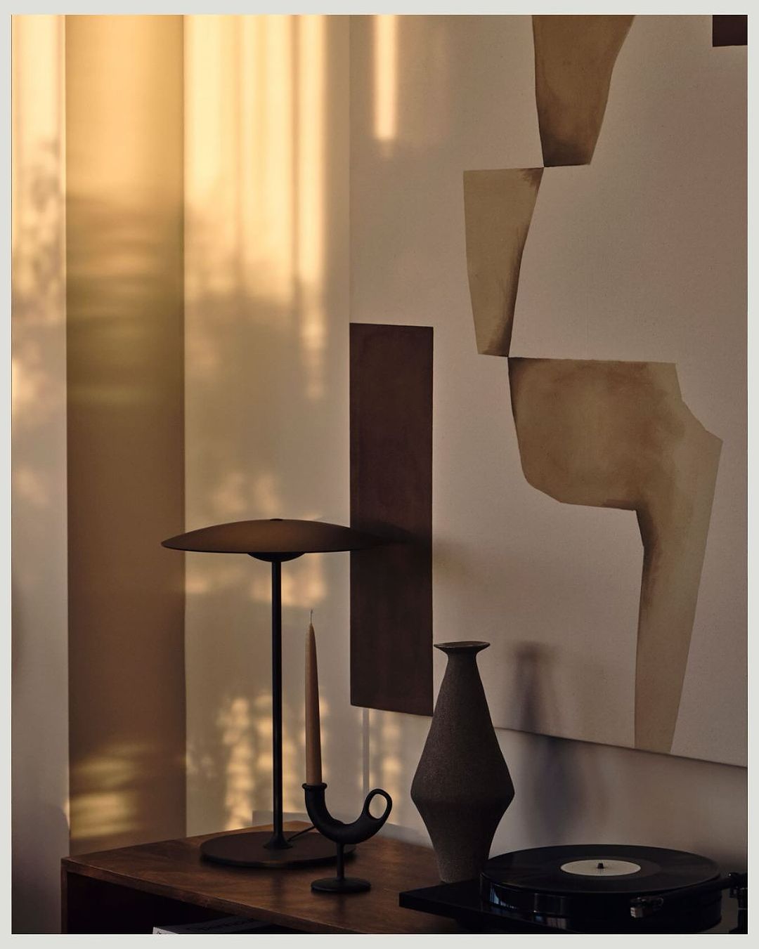 Still life photograph of a lamp, candle holder with candle, vase and record player on a wooden table against neutral walls.