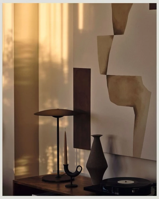Still life photograph of a lamp, candle holder with candle, vase and record player on a wooden table against neutral walls.