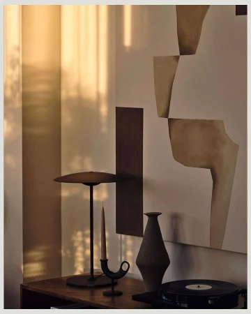 Still life photograph of a lamp, candle holder with candle, vase and record player on a wooden table against neutral walls.