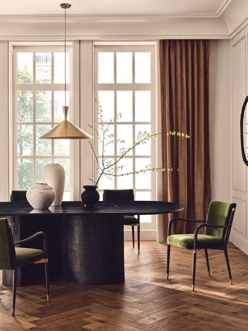 Dining room interior with a black dining table, green chairs, pendant lighting, hardwood floors, vases, and decorative tree branches.