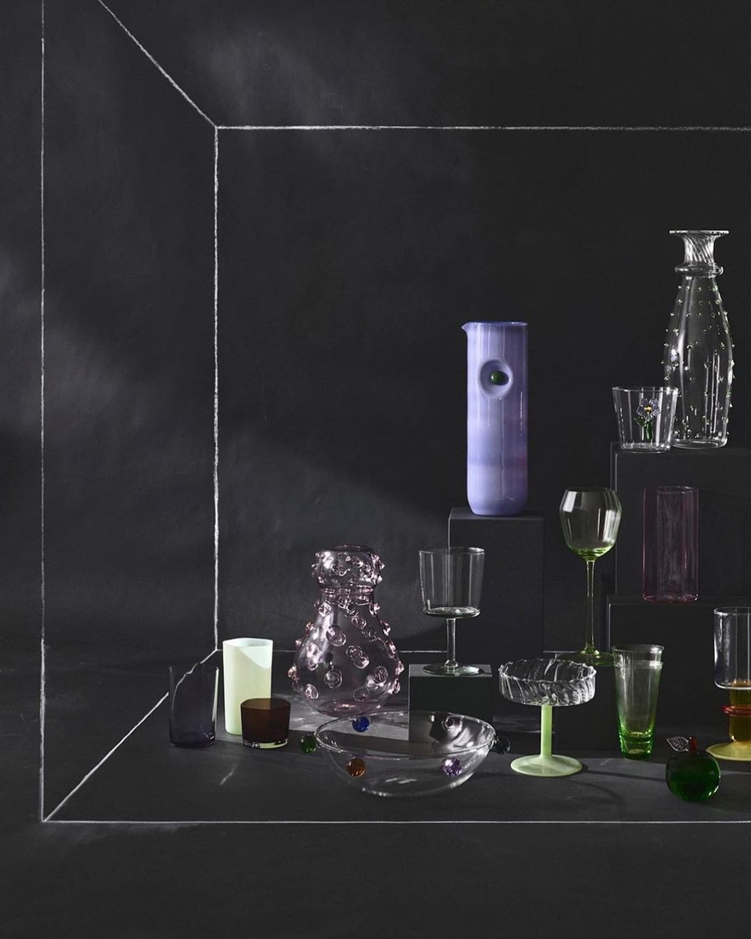 Still life shot of a curated assortment of decorative glassware in an arrangement featuring clear, green, and purple tones against a dark backdrop.