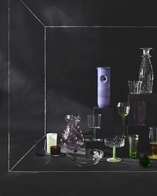 Still life shot of a curated assortment of decorative glassware in an arrangement featuring clear, green, and purple tones against a dark backdrop.