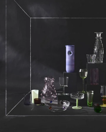 Still life shot of a curated assortment of decorative glassware in an arrangement featuring clear, green, and purple tones against a dark backdrop.