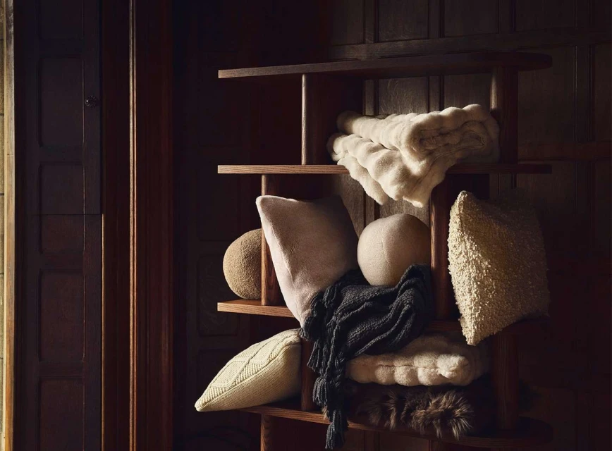 Cozy home decor featuring textured throw blankets, decorative pillows, and a soft wool scarf arranged on a wooden shelving unit.