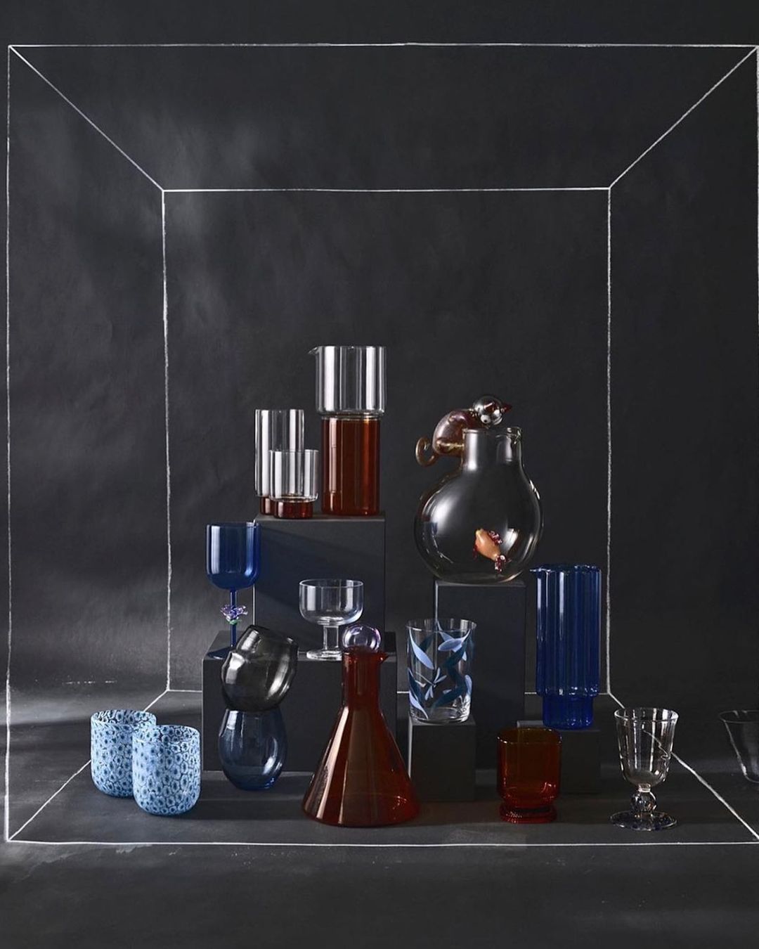 A collection of clear glass drinkware in blue, red, and clear tones is arranged on pedestals inside a sketched box in a studio.