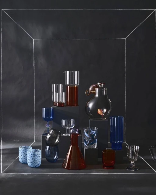 A collection of clear glass drinkware in blue, red, and clear tones is arranged on pedestals inside a sketched box in a studio.