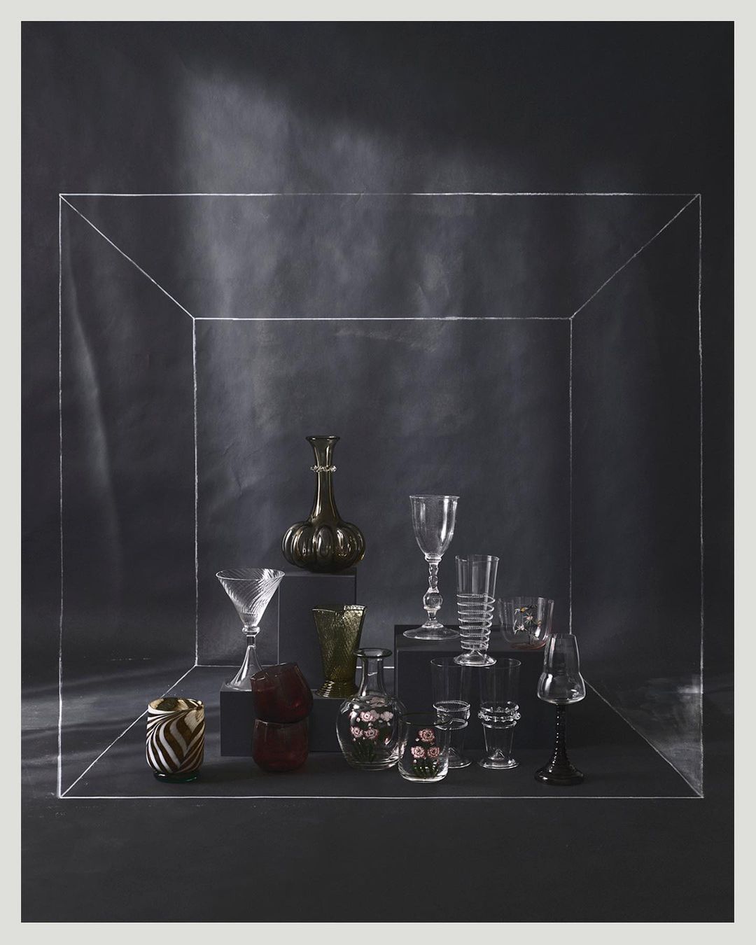 Elegant still life featuring an assortment of glassware displayed on dark platforms against a deep grey backdrop within a clear framed box.