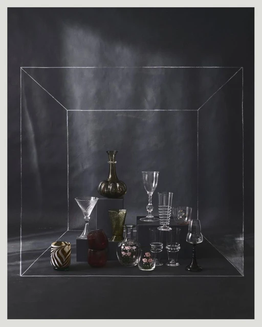 Elegant still life featuring an assortment of glassware displayed on dark platforms against a deep grey backdrop within a clear framed box.