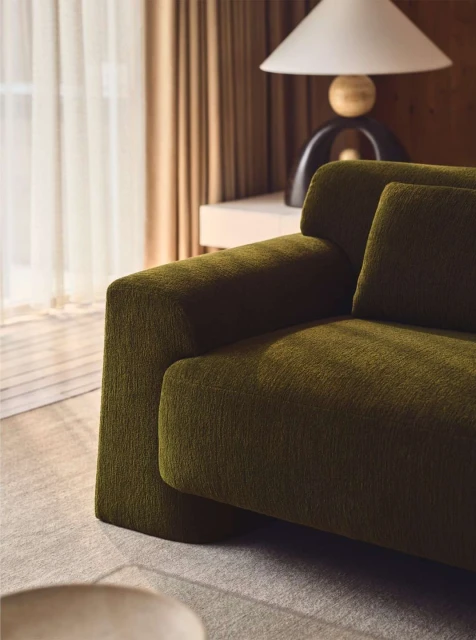 Olive green sofa with throw pillow against light beige wall with sheer curtains, side table, and modern lamp in home interior.