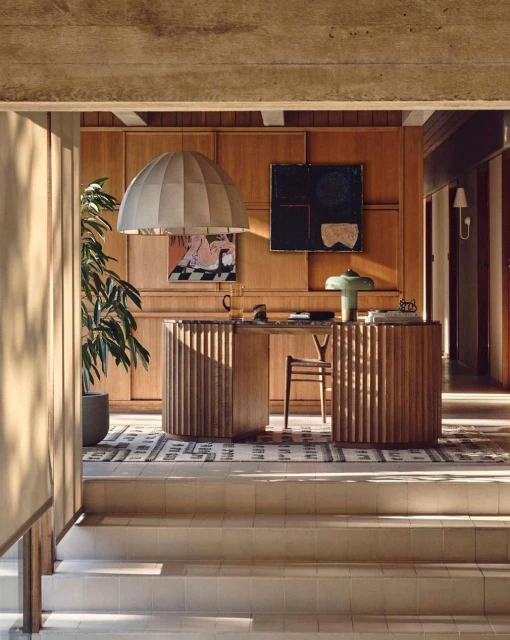 Interior view of home office with geometric rug, wood paneled walls, two desks, pendant light, indoor plant, and art on the walls.