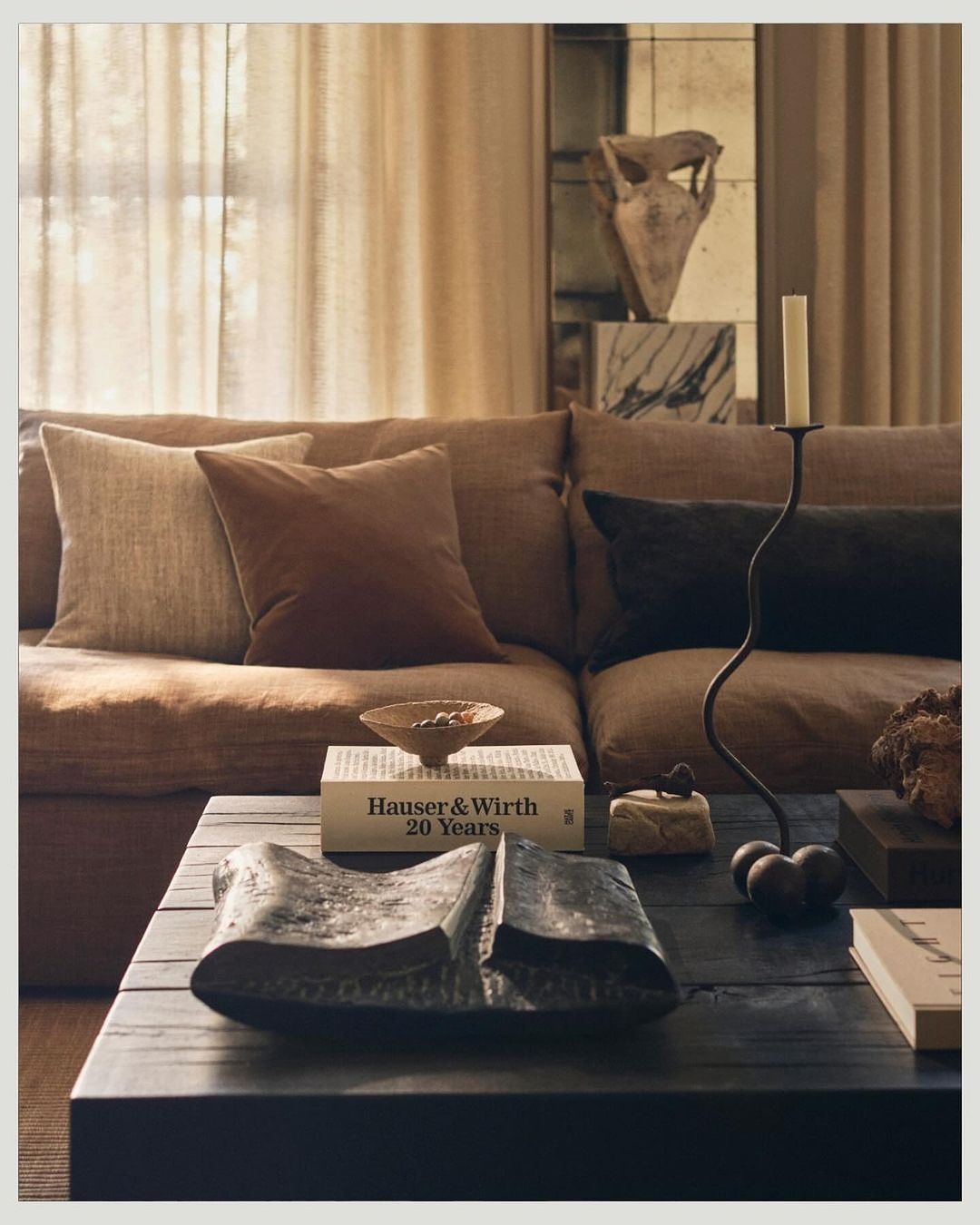 Living room interior with a brown couch, assorted cushions, dark coffee table with decorative objects, and beige drapes in a neutral color palette.