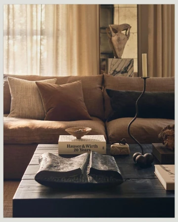 Living room interior with a brown couch, assorted cushions, dark coffee table with decorative objects, and beige drapes in a neutral color palette.