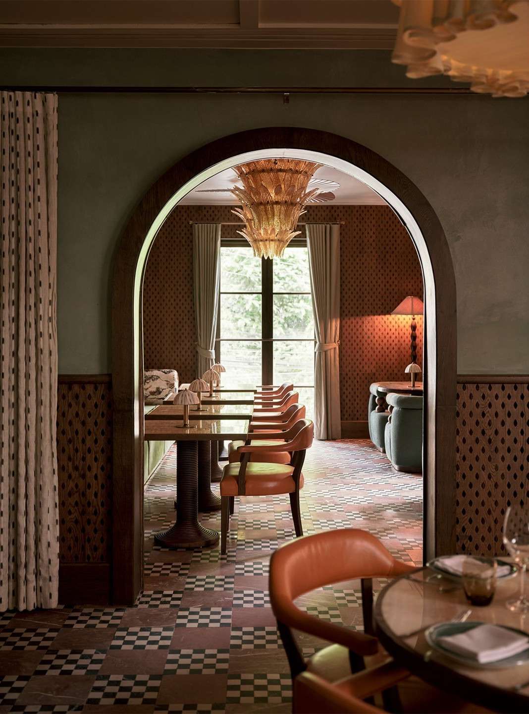 Ornate restaurant interior featuring tables with orange chairs, an archway, checkered floors, patterned walls, and a hanging chandelier.