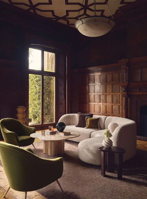 Cozy living room interior with a white boucle sectional sofa, olive green armchairs, a marble coffee table, and dark wood paneling.