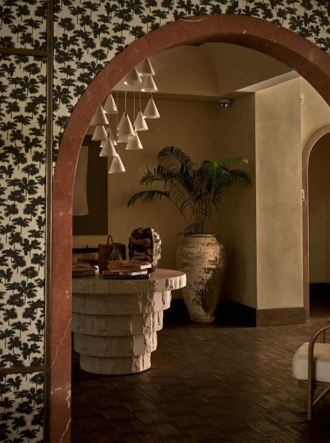 View through an archway to an interior with a textured round table, books, bags, white pendant lights, and a potted plant.