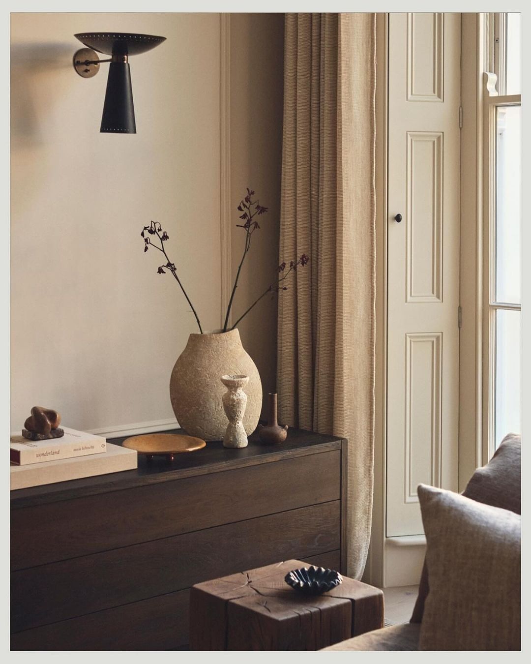 Interior still life with a beige textured vase holding decorative branches, and a dark wood dresser with books and decorative objects.