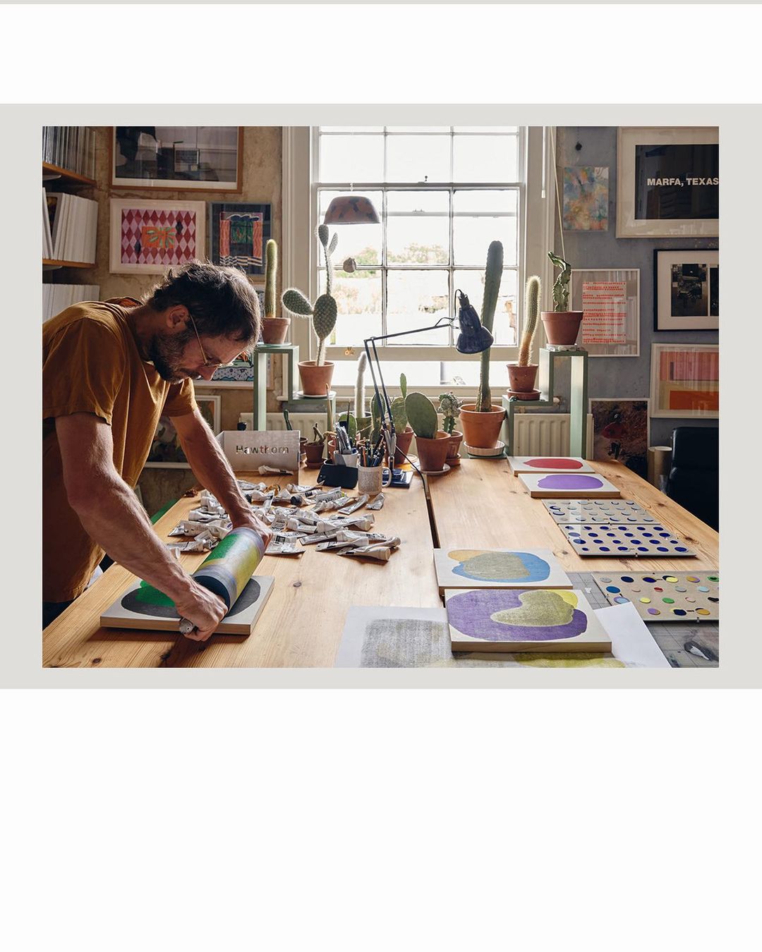 Artist in his studio using a roller to apply paint to a canvas. A long table is covered with art supplies and cacti in pots.