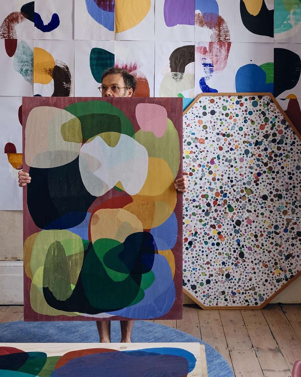 Artist holding abstract painting with colorful organic shapes in a studio setting, surrounded by other artworks and white walls.