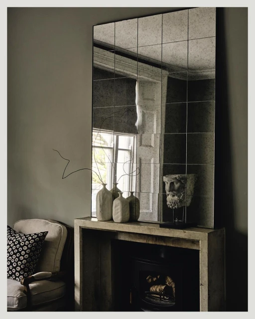 Interior photo: Modern living room featuring a mantelpiece with vases, head bust, a framed mirror, an armchair and neutral wall color.