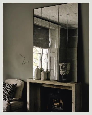 Interior photo: Modern living room featuring a mantelpiece with vases, head bust, a framed mirror, an armchair and neutral wall color.