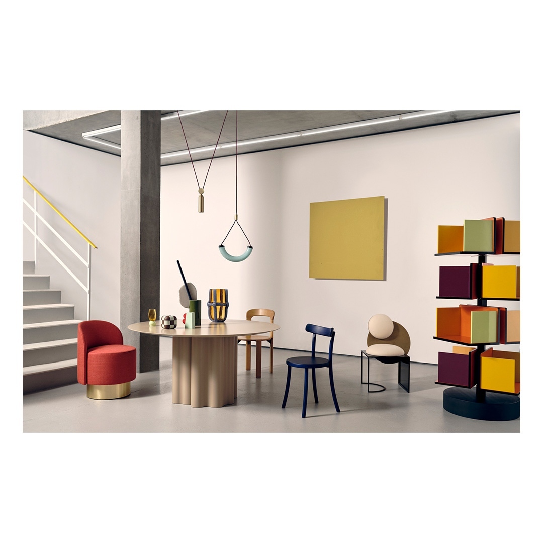 Interior design photo showcases a round table surrounded by colorful chairs in a room with modern, minimalist decor and a yellow wall art.