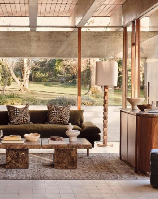 Living room interior with a brown sofa with patterned pillows, a stone coffee table, floor lamp, and view of a green lawn and trees.