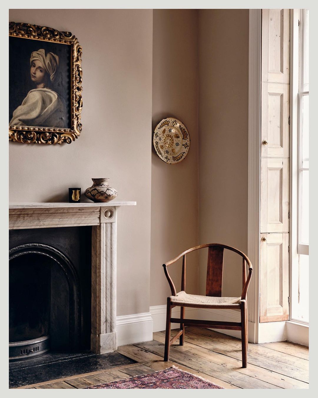 Interior design featuring a vintage painting, fireplace, wooden chair, and decorative dish in a room with light brown walls and wood floor.